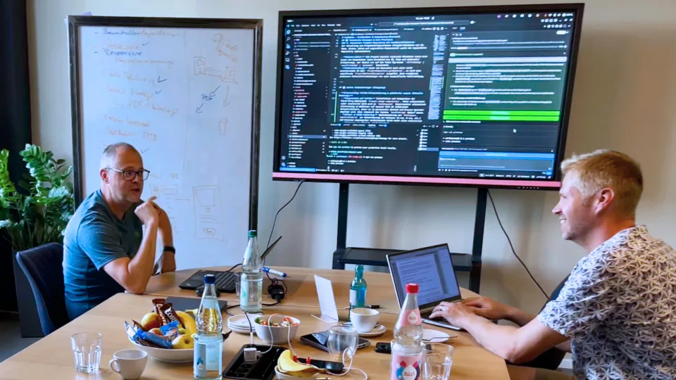 Zwei Personen arbeiten im Co-Creation Workshop am Whiteboard und am Code auf dem Wandbildschirm